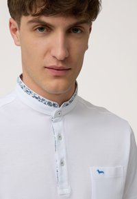 Hvid polo-shirt med trykt blåt paisley-mønster indeni kraven, korteærmer og en lille blå emblem på brystlommen.