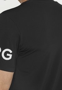 Camisa atlética negra con mangas cortas, textura suave y letras blancas con puntos en la manga izquierda. Diseño simple con cuello redondo.