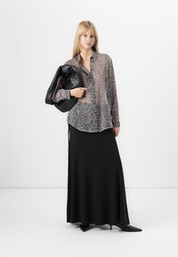 Mulher em pé, vestindo uma blusa transparente de padrão de leopardo, saia longa preta, sapatos de salto pretos e agudos, segurando uma grande bolsa de couro preta.