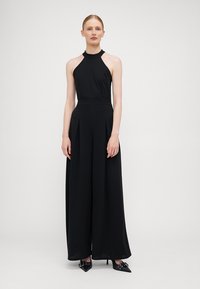 Black halter jumpsuit with a fitted bodice and wide-legged trousers. Features pleats and a smooth, textured fabric. Elegantly simple design.