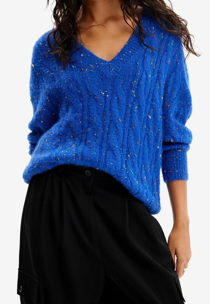 Femme portant un pull en maille torsadée bleu vif moucheté à col en V avec manches longues rentré dans un pantalon noir ample.