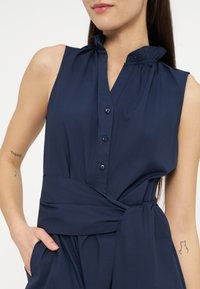 Γυναίκα που φορά αμάνικο μπλε σκούρο φόρεμα με κουμπιά, βολάν στον γιακά και φαρδύ υφασμάτινο ζωνάρι, με το χέρι στην τσέπη, τατουάζ σε και τα δύο χέρια.