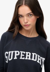 Marineblaue Sweatshirt mit gerippter Textur und weißer "SUPERDRY" Aufschrift über der Brust; verfügt über Raglanärmel und ein Rundhals-Design.
