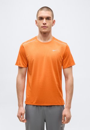 Ung mand står oprejst iført orange Nike t-shirt med korte ærmer og grå shorts mod en ensfarvet lys baggrund.