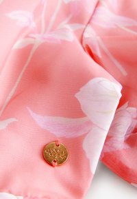 Svetlo roza tkanina z cvetličnimi vzorci, s okroglim gumbom iz zlatega kovine z vgraviranim cvetličnim vzorcem, pritrjenim z oranžno nitjo.