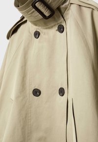 Béžový trenčkot s dvojradovými tmavými gombíkmi, vysokým golierom so zapínaním na pracku a záhybovým detailom na boku.