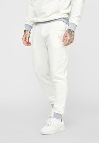 Krämfärgade joggers med grå resår, med en enkel design och liten präglad logotyp. Kombineras med vita sneakers. Slät tygtextur.