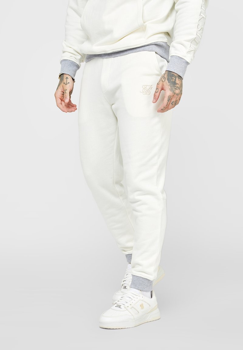 Krämfärgade joggers med grå resår, med en enkel design och liten präglad logotyp. Kombineras med vita sneakers. Slät tygtextur.