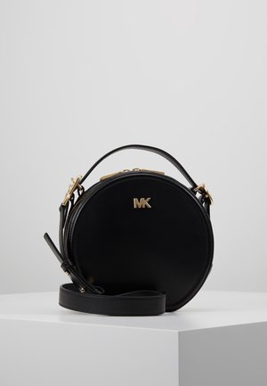 Schwarze runde Ledertasche mit goldenem MK-Logo, oberem Griff und abnehmbarem Schulterriemen auf weißem Podest präsentiert.