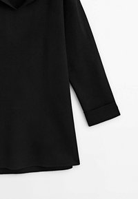 Chemise noire à manches longues avec poignets repliés, vue de la taille aux épaules sur fond blanc.