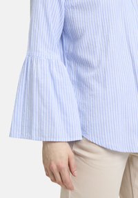 Hellblau gestreifte Bluse mit Glockenärmeln und strukturiertem Stoff. Abgerundeter Saum und dezenter Glanz des Materials.