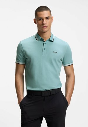 Man draagt lichtblauw poloshirt met korte mouwen en kraag, en zwarte broek, staat met handen in de zakken tegen een witte achtergrond.