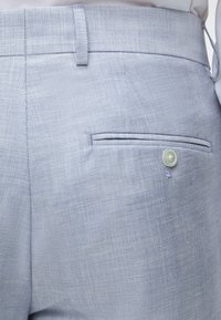 Pantalon de costume bleu clair texturé avec une poche arrière boutonnée et des passants pour ceinture, porté sur une chemise blanche.