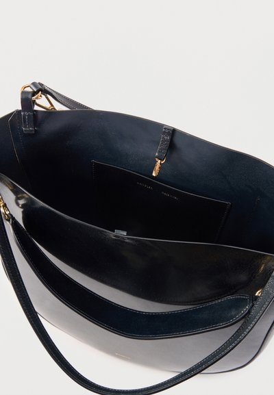 Μαύρη δερμάτινη τσάντα χειρός, ανοιχτή για να φαίνεται η άδεια εσωτερική τσέπη με χρυσό κούμπωμα και ιμάντα ώμου σε απλό φόντο.