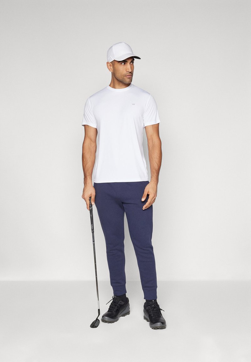 Bílý tričko s krátkým rukávem v kombinaci s tmavě modrými tepláky a černými golfovými botami; drží golfovou hůl; má na sobě bílou čepici.