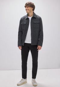 Giacca grigia in misto lana con colletto, due tasche sul petto e chiusura con bottoni, abbinata a una maglietta bianca e pantaloni neri. Sneakers bianche.