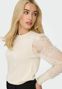 Cremefarvet pullover med lange, gennemsigtige ærmer prydet med rynkede stofdetaljer. Tæt siddende design med rund halsudskæring og ribbede manchetter.