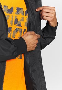 Sort lynlåsjakke med en tekstureret overflade, båret over en orange skjorte med grafisk design i marineblå og olivengrøn.