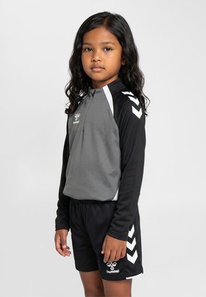 Ung pige med langt hår iført sort og gråt sportstøj med hummel-logoer og hvide Chevron-mønstre på ærmer og shorts.