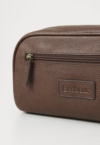 Pouch de couro texturizado castanho com fecho zip na parte superior e patch do logótipo Barbour na frente, apresentado contra um fundo claro e liso.