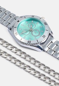Relógio de aço inoxidável prateado com um mostrador verde, apresentando três submostradores, bracelete ligado e uma corrente simples ao lado.