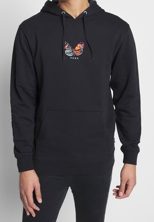 Mann trägt schwarzen Hoodie mit buntem Schmetterling und "VANS"-Logo auf der Brust sowie schwarze Hose, steht vor einem einfarbigen Hintergrund.