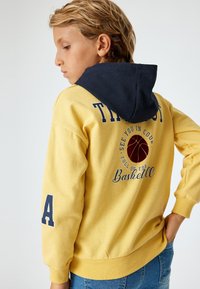 Sudadera amarilla con capucha azul marino, que presenta un gráfico de baloncesto y texto en la parte posterior. Tejido suave, puños acanalados y un corte relajado.