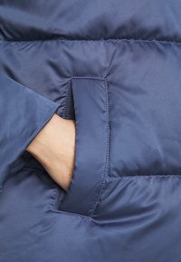 Granatowa kurtka puchowa o gładkiej, błyszczącej fakturze, z boczną kieszenią o prostokątnym otworze i kontrastującymi szwami.