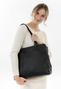 Sac en cuir noir avec une finition texturée et deux poignées robustes, tenu par un mannequin portant une robe légère côtelée.