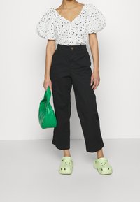 Blusa bianca con motivo a cuori neri, maniche a sbuffo; pantaloni neri a gamba larga; borsa verde testurizzata; zoccoli verde chiaro con fori di ventilazione.