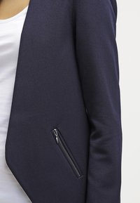 Námořnický blazer se strukturovaným designem, vyrobený z texturované tkaniny. Obsahuje zipovou kapsu na boku a čistou, upravenou siluetu.