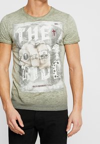 Grön T-shirt med kort ärm som har en grafisk design med skallar och text, tillverkad av mjukt tyg med en blekt struktur. Svarta byxor bärs under.