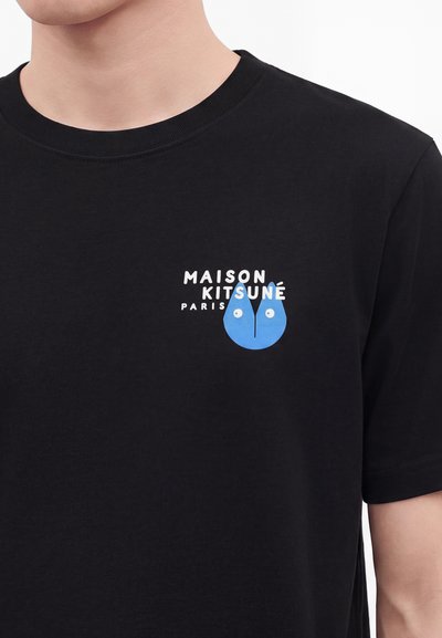 Μαύρο t-shirt με κείμενο "Maison Kitsuné Paris" και μπλε αφηρημένο λογότυπο προσώπου αλεπούς στο αριστερό στήθος, φορεμένο από πρόσωπο περικομμένο στον λαιμό.