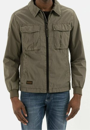 Mann trägt eine olivgrüne Jacke mit Reißverschluss, zwei Brusttaschen mit Klappen und elastischen Bündchen, kombiniert mit blauen Jeans.