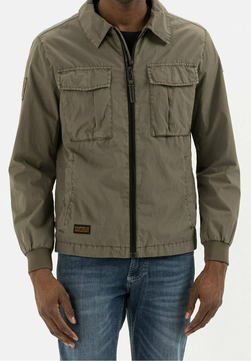 Homme portant une veste zippée vert olive avec deux poches poitrine à rabat et des poignets élastiques, associée à un jean bleu.