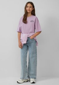 Lila oversized t-shirt met grafische tekst, gedragen over lichtblauwe wijde jeans. Vergezeld van een lichte trui om de taille gebonden en witte sneakers.
