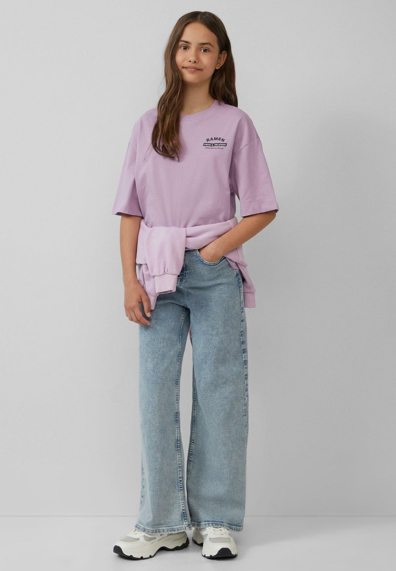 Lila oversized t-shirt met grafische tekst, gedragen over lichtblauwe wijde jeans. Vergezeld van een lichte trui om de taille gebonden en witte sneakers.