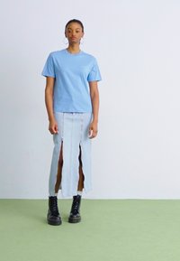 T-shirt in cotone azzurro abbinato a una gonna di jeans con spacchi frontali, completato da stivali neri con lacci su un pavimento verde chiaro.