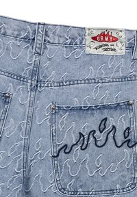 Vaqueros de mezclilla azul con un patrón de olas cosido en blanco, con un diseño de llama negra en el bolsillo trasero y una etiqueta de marca en la cintura.