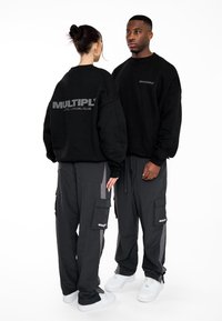 Černé oversized mikiny s nápisem "MULTIPLY" vytištěným na zadní straně; v kombinaci se šedými cargo kalhotami s bočními kapsami a bílými teniskami.