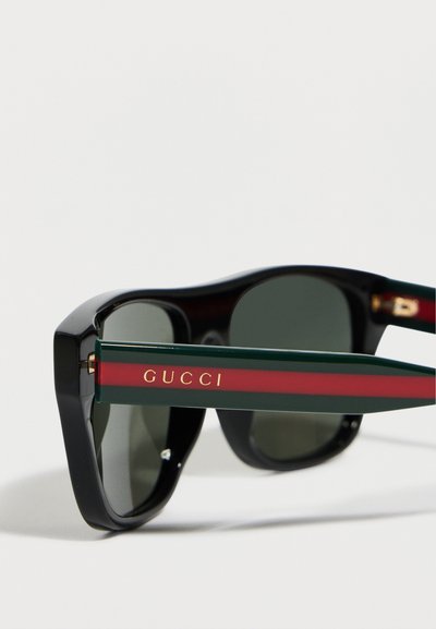 Μαύρα γυαλιά ηλίου με σκουρόχρωμους φακούς και πράσινα-κόκκινα μπράτσα που φέρουν το χρυσό λογότυπο «GUCCI» στο πλάι.