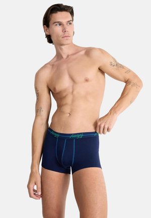 Oberkörperfreier Mann mit Tätowierungen, der dunkelblaue Boxershorts mit grünem "Sloggi"-Bund richtet und seitlich gegen einen schlichten Hintergrund blickt.