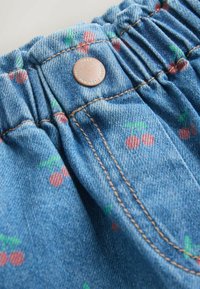 Prenda de mezclilla con cintura fruncida, de color azul claro y pequeños estampados de cerezas en rojo y verde. Incluye detalles de herrajes de metal.