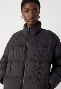 Svart polstret jakke med quiltede seksjoner, høy krage og glidelås; laget av lett, teksturert stoff med en liten logodetalj.