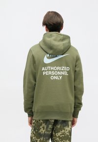 Õlivaroheline kapuutsiga sviiter, millel on ees suur tasku ja seljal suur helesinine Nike logo koos tekstiga "AINULT VOLITATUD ISIKUTELE".