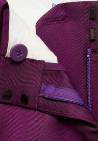 Tissu texturé violet avec une doublure intérieure blanche. Comprend un bouton violet, des accents noirs et un détail de poche zippée visible.