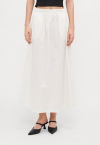Jupe maxi blanche avec une taille élastique, présentant une texture lisse et un design fluide. Associée à des talons noirs à bout pointu.