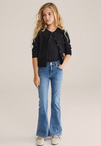 Schwarze Strickjacke mit goldenen Knöpfen, enges schwarzes Oberteil, ausgestellt helle blaue Jeans und hellgrüne Schuhe. Das Model hat lange, wellige Haare.