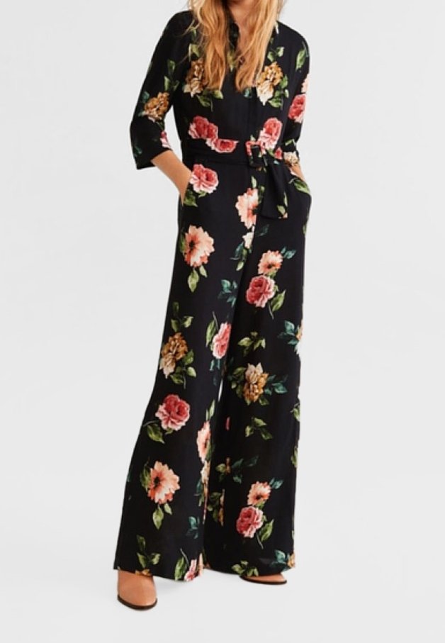 Femme portant une combinaison noire à motif floral avec jambes larges, manches trois-quarts et poches avant, associée à des talons couleur camel.
