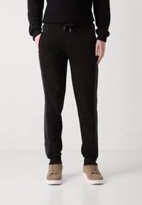 Pantalones de chándal negros con una cintura ajustable por cordón, rayas laterales y un panel texturizado a lo largo de las piernas; combinados con zapatillas en tono beige.